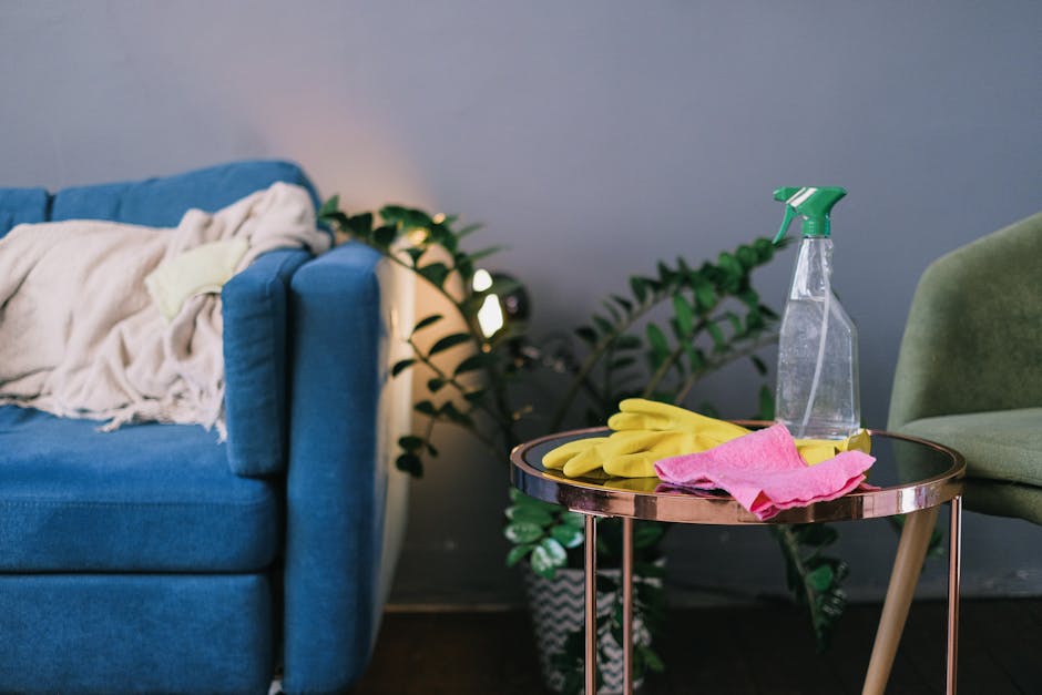 A blue fabric sofa with a cream blanket draped over one armrest, situated in a living room corner. Next to the sofa, a small round side table with a reflective surface holds yellow cleaning gloves, a 