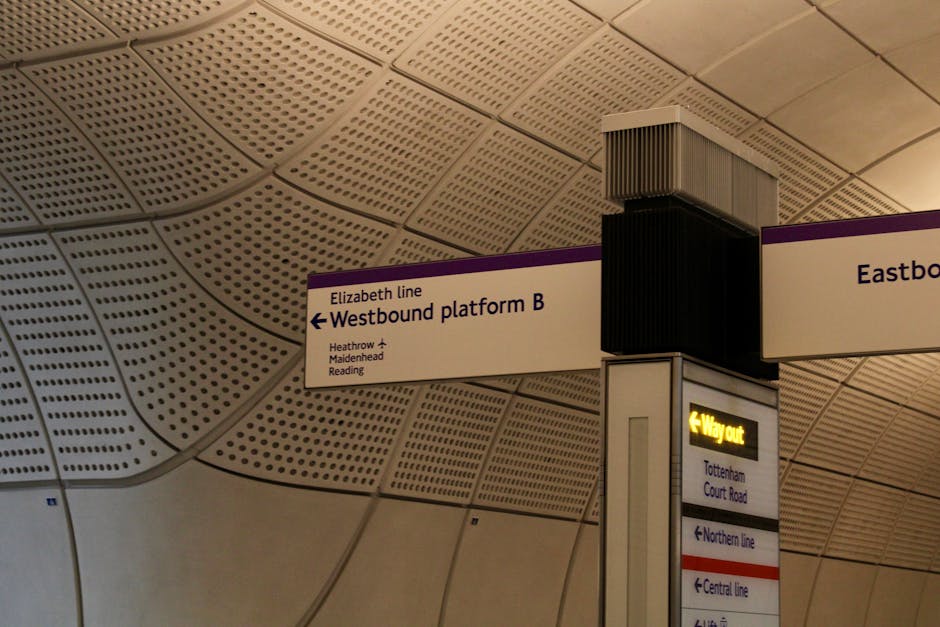 Interior view of a London Underground station ceiling featuring curved beige acoustic panels with small perforations. A vertical column displays directional signage, including a white sign with purple